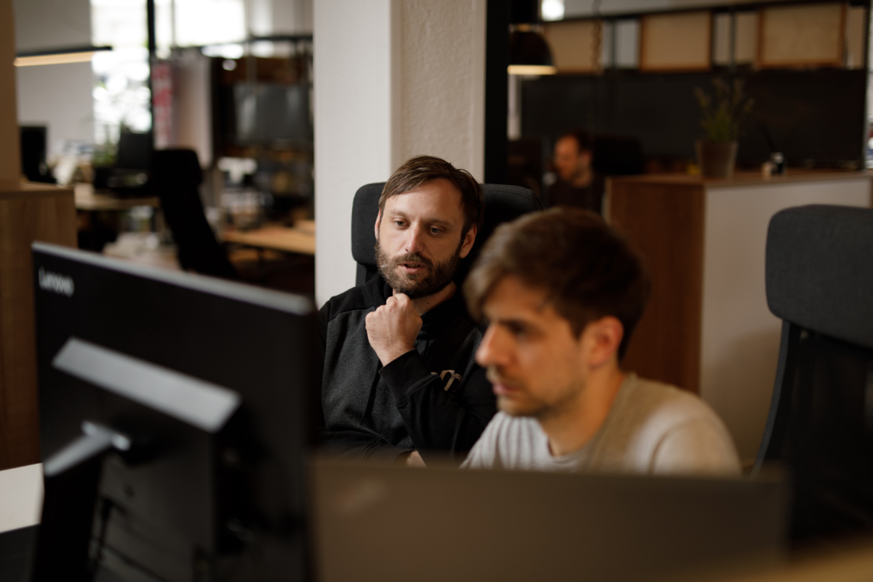 Zwie Mitarbeitende nebeneinander im Gespräch an ihrem Arbeitsplatz.