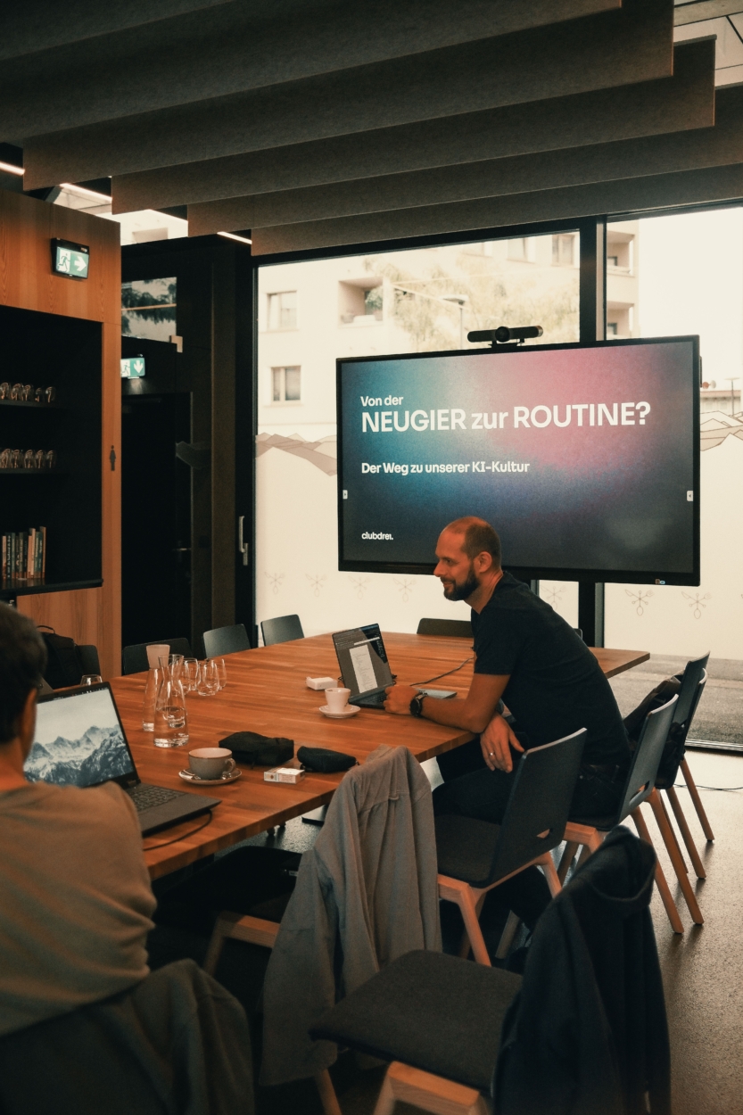 Ein Mitarbeiter an einem Besprechungstisch. Im Hintergrund steht auf einem großen Bildschirm "Von der Neugier zur Routine. Der Weg zu unserer KI-Kultur".