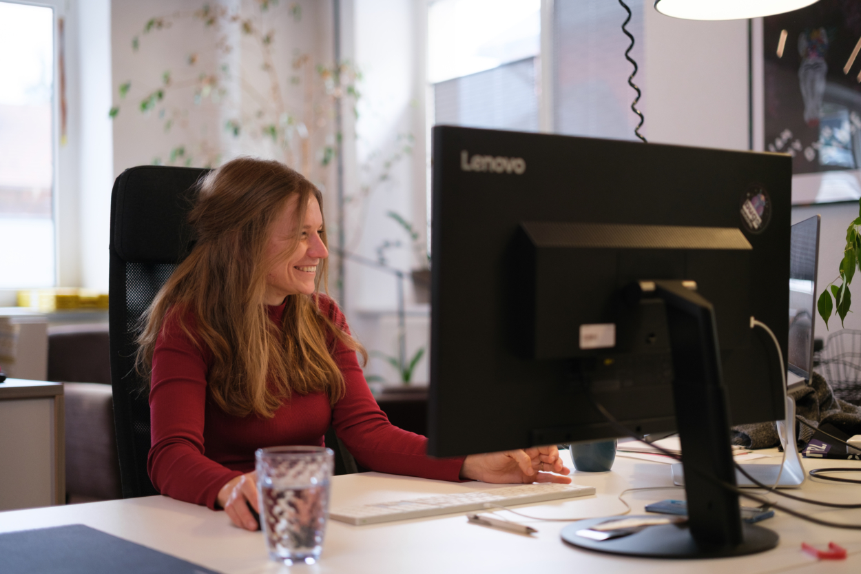 Eine Mitarbeiterin in einem roten Pulli sitzt an ihrem Arbeitsplatz und lächelt.