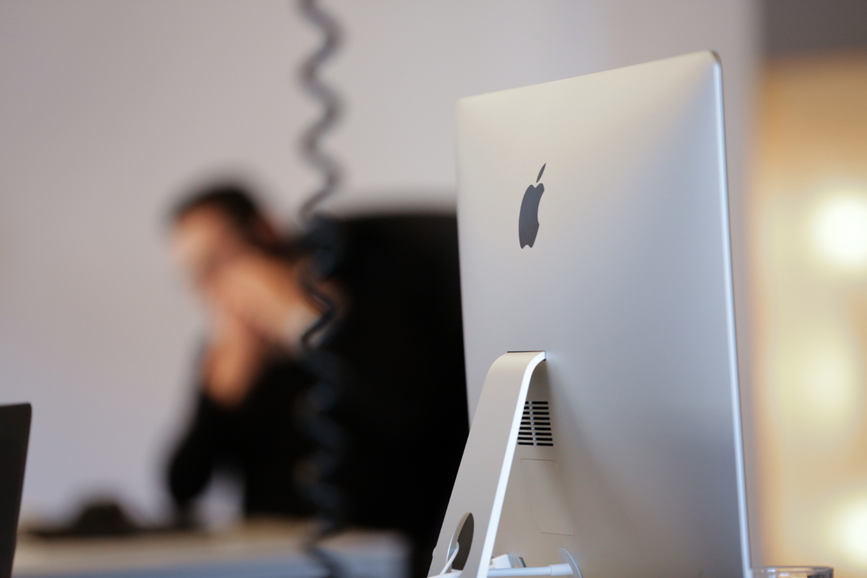 Ein Bildschirm. Im Hintergrund ist ein Miterabeiter der telefoniert.